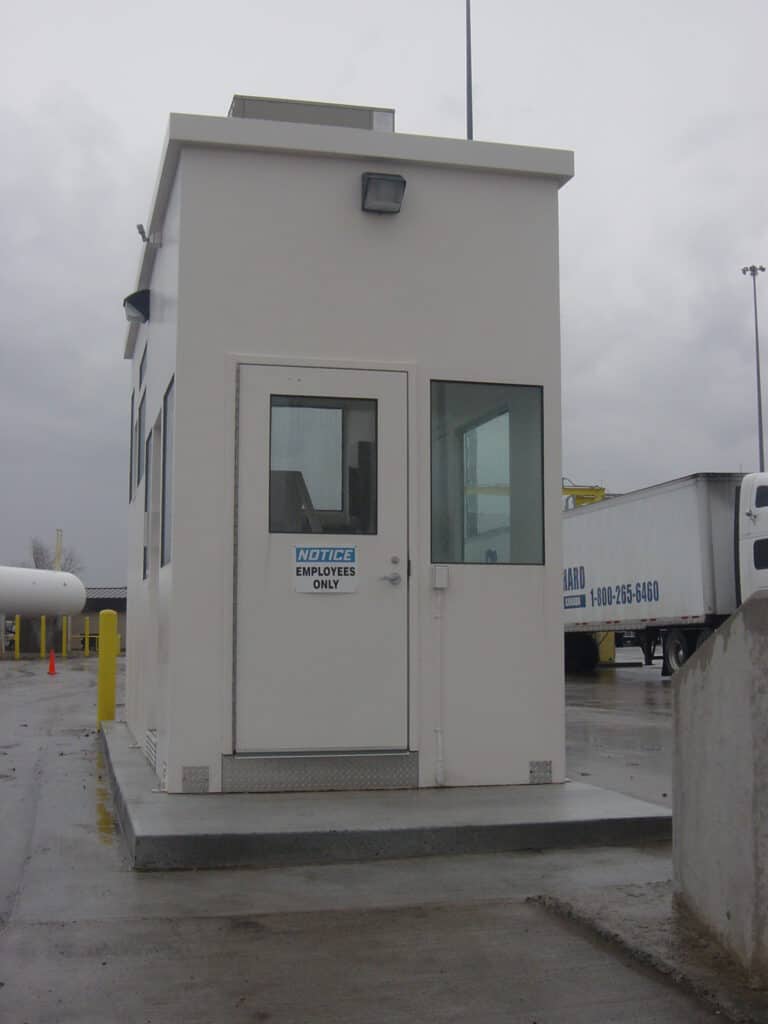 A small, square security booth with a white exterior, labeled "employees only", features a ballistic rated door and is situated at an industrial entrance with trucks and concrete barriers visible.