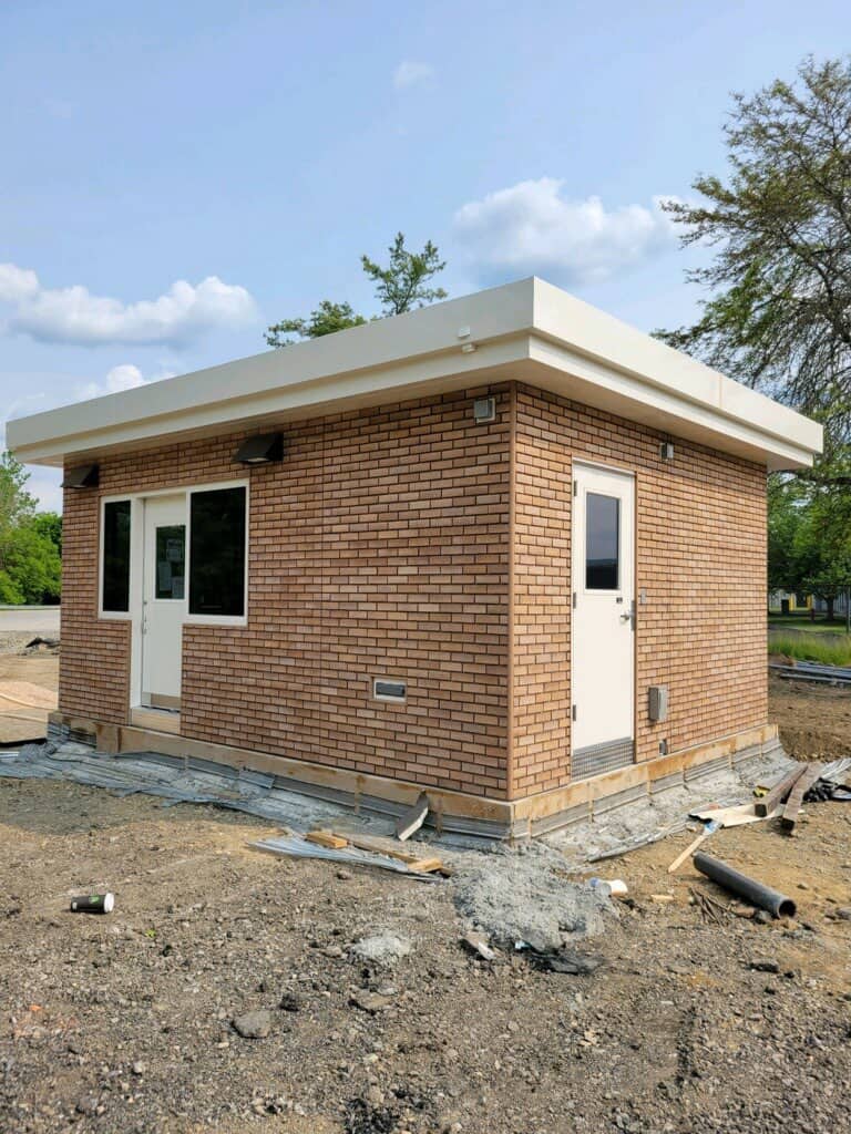 A small, new building with factory-installed brick and a flat roof, featuring a white door and two windows, located on a construction site with scattered debris around.