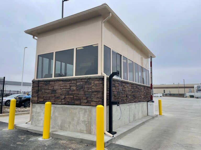 Small beige security booth with large windows and Factory Installed Faux Stone paneling at its base, located in a parking lot with yellow bollards, under an overcast sky.