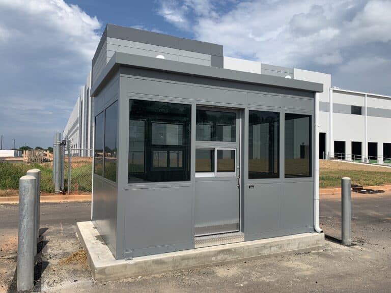 A new small gray modular building with large windows and a standard double door, located in a developing area with concrete bollards in front and an industrial building in the background under a cloudy sky.