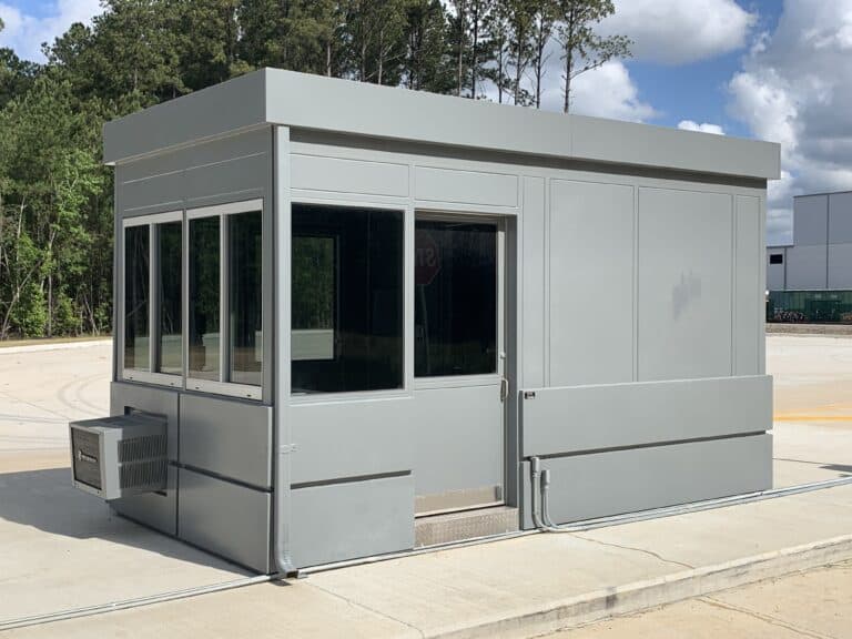 A small, modern gray security booth with a steel sliding door and large windows, situated on a concrete surface under a clear sky. The booth is equipped with an air conditioning unit.