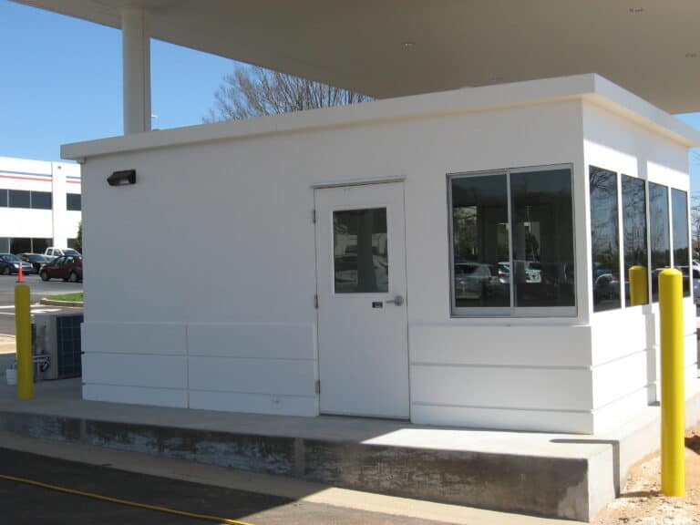 A small, white security booth with a flat roof, featuring a steel swing door and a window, stationed in a parking lot under a clear blue sky. Yellow bollards are visible around the booth