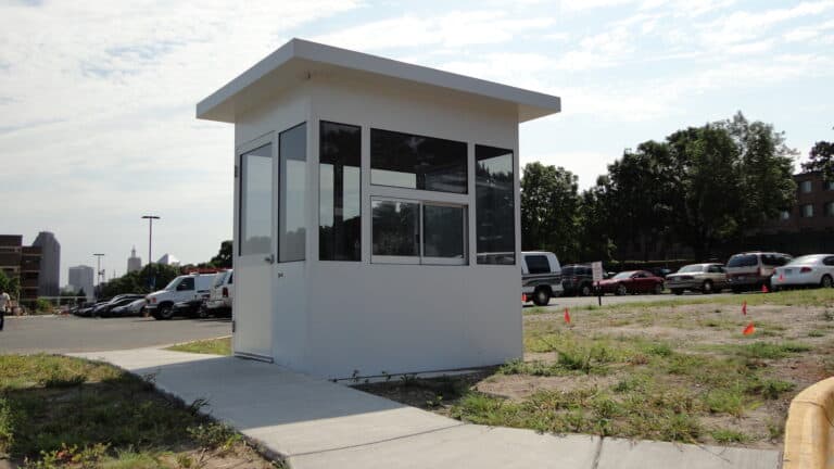 A small white security booth with a transaction window, situated on a concrete path at the edge of a parking lot, with a city skyline in the background and clear blue sky.