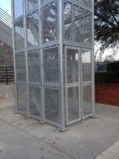 A metal outdoor elevator shaft made of gray metal grills, located next to a stone building, with a glimpse of greenery and red fencing in the background.