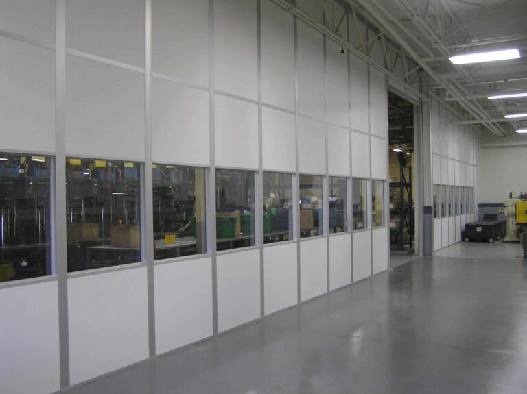 A spacious industrial facility with large glass windows through which machinery and workstations are visible. The interior, enhanced with acoustic solutions, is brightly lit, reflecting off the white-tiled floor.
