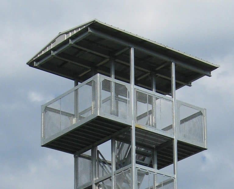 A metal fire lookout tower with a flat roof and protective railings, situated against a cloudy sky in an urban high-rise setting.