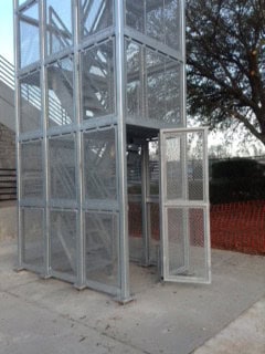 An outdoor metal elevator shaft with mesh security door panels and an open gate, surrounded by trees and a building in the background.