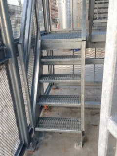 Metal staircase with diamond pattern steps, enclosed by mesh and metal railings, in an outdoor industrial setting.