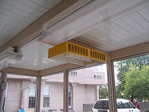 A photo showing an electric heater installed in the ceiling corner of a building's entrance, featuring a yellow casing and black grid, with visible surroundings of glass doors and windows.