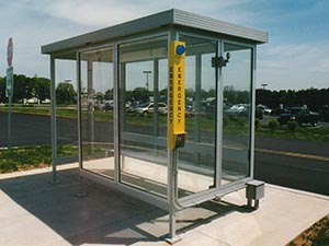 A clear bus shelter with a glass structure and a metal roof installed on a concrete pad, featuring an emergency call box with electronic components and a blue light, located in a sunny parking area.