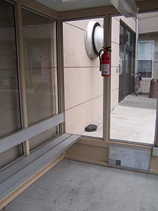 An interior view of a smoking area with glass walls, featuring aluminum benches, a wall-mounted ashtray, and a fire extinguisher. Transition to outdoor space is visible.