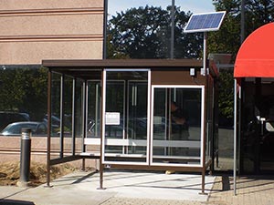 A modern bus shelter with glass walls and doors, and a solar panel battery installed on the roof, located next to a building under a bright sky.