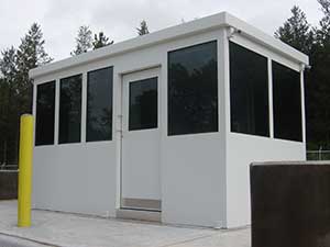 A small, modern white security booth with large windows on three sides and a ballistic rated sliding door, located in a parking area near a forest. A yellow bollard is visible on the left side