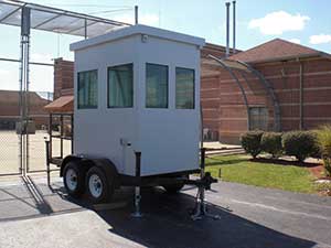 A portable security booth on a trailer parked outside a building under a clear sky features large windows, stabilizers, and a standard roof.