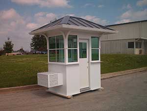 A small, portable security booth with a white exterior and gray extended roof, featuring multiple windows, located on a concrete surface against a clouded sky and grassy background.
