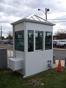 A small security booth with multiple glass windows and a hip roof, located near a street, equipped with an external air conditioning unit and a streetlight overhead.