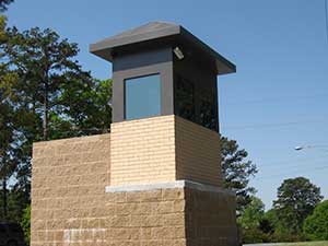 A small, modern security guard booth made of brick with expanded metal mesh windows and a flat roof, situated under a clear blue sky with trees in the background.