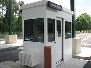 A small, modern security booth with standard wall construction and black trim is situated in a parking area, featuring large windows and an air conditioning unit on top.