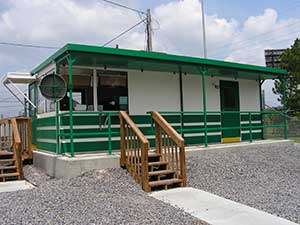 A small green and white building with ballistic rated walls and a flat roof, featuring a front porch, wooden stairs, and surrounded by a gravel area.