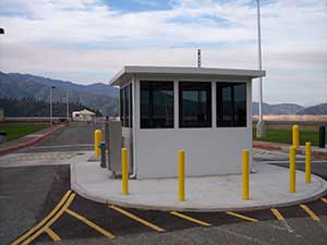 A small security guard booth at the entrance of a facility, surrounded by yellow bollards and parking lines, with ballistic rated fixed windows and mountains in the background.