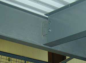 Close-up of a construction beam connection featuring bolts and plates, part of a building's structural framework, against a yellow wall background.