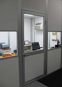 An office doorway, designed as a single man door of heavy duty aluminum, reveals a glimpse into a small room with gray walls, equipped with a chair, desk, computer, and a wall-mounted air