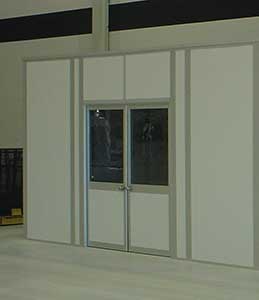 A set of double doors with glass panes, crafted from heavy-duty aluminum, is set in a wall with a light gray color. A dim reflection of a person is visible in each glass pane.