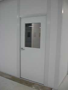 A modern white steel door with a rectangular glass window set in a clean, white wall in a well-lit interior space. The floor is light gray.