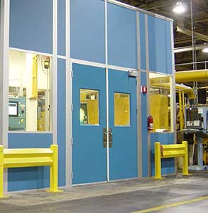 A well-lit industrial workspace with a large blue partition wall featuring windows and yellow safety guards. There are steel double doors in the center, leading to another section of the facility.