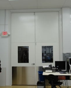 Office room with a closed steel double door and two small windows. There is a computer desk with a monitor in the foreground. A bright environment with neutral colors.