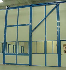 A large custom industrial sliding door with a blue metal frame and multiple glass panels, mounted in a light-colored wall inside a spacious room.