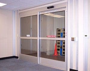 Automated sliding glass doors in an office building, showing a glimpse of a brightly lit storage room with shelves and a traffic cone inside.