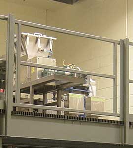 Industrial machinery in a factory setting with visible safety barriers, including a 6' gate, featuring a conveyor system and various mechanical components.