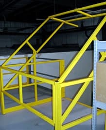 Yellow metal livestock gate resembling a geometric structure inside a warehouse, with a background of grey floors and partial views of shelving units.