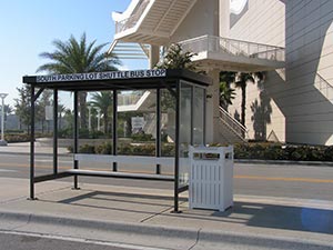 A compact, stylish bus stop shelter featuring a black metal frame and a transparent back panel, situated on an urban street alongside palm trees and a white, futuristic building. The sign displays "Borrego