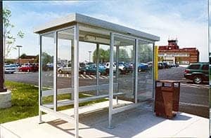 A simple, style B bus shelter with a metal frame and glass walls, located in a sunny parking lot with various cars and a building in the background.