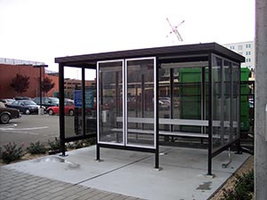 A modern Style C bus shelter with a glass enclosure and a metal frame, located on a paved area next to a parking lot and green dumpsters, under an overcast sky.