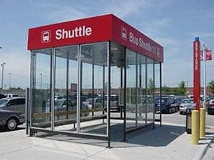 A modern glass-walled shuttle bus stop with a red "shuttle" sign on top and a raised fascia, located in a parking area under a clear blue sky.