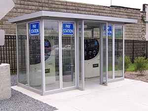 Three pay stations housed in a small, modern glass enclosure with a standard flat roof, situated on a concrete pad next to a stone wall. Signs on the glass panels read "Pay Station.