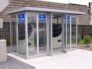Three pay stations housed in a small, modern glass enclosure with a standard flat roof, situated on a concrete pad next to a stone wall. Signs on the glass panels read "Pay Station.