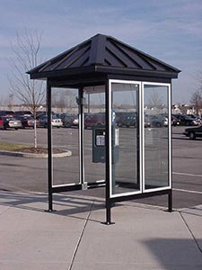 A small, modern bus shelter with glass walls and a standing seam hip roof, located on a concrete sidewalk beside a parking lot under a clear sky.