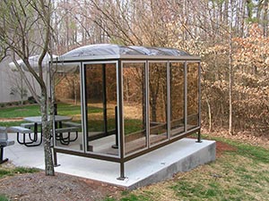 A metal and glass bus shelter with an acrylic dome roof, located on a concrete slab in a park, surrounded by trees and a picnic table nearby.