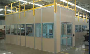 A modern cleanroom with glass walls and a single swing door located inside a large industrial facility, under a yellow overhead structure.