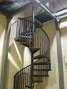 A black metal spiral staircase inside a building with beige walls and a gray metal railing, featuring an innovative stair design, leads to a higher floor.