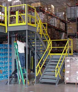 A warehouse interior featuring a custom staircase in yellow leading up to a platform, with a man on a ladder and various supplies, including blue crates and cardboard boxes, organized on shelves.