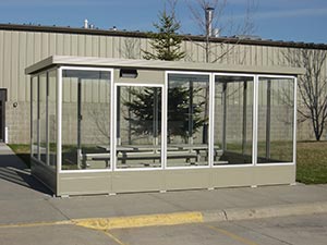 A small, modern smoking shelter with enclosed lower panel glass walls and a grey frame, featuring benches and a table inside, located next to a building in a paved area.
