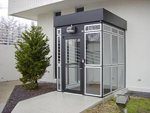 A modern, small full height glass and metal smoking shelter with a sliding door, positioned next to a building and a green bush, under an overcast sky.