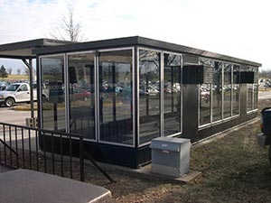 A full height solid-paneled bus shelter with a bench inside, situated by a parking lot with cars and trees in the background under a clear sky.