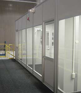 An image of a clean, modern interior featuring a glass-walled office with transparent glass panels and aluminum frames, set against the backdrop of a gray carpeted floor and industrial walls.