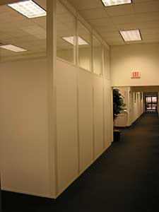 Interior of an office with partition walls featuring top of panel glazing, fluorescent ceiling lights, a dark carpeted floor, and a visible entrance door in the distance.